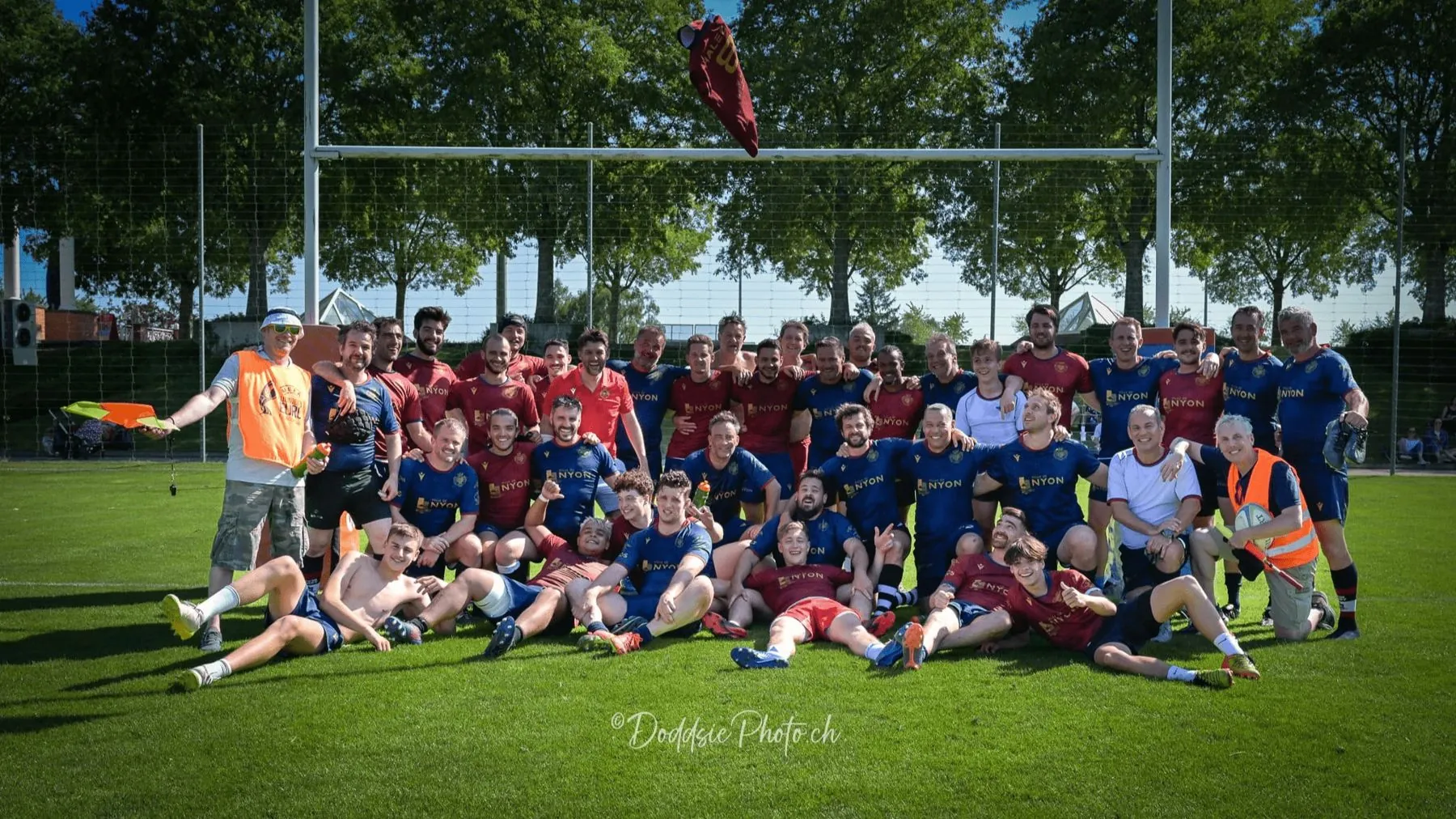 Photo de groupe : les joueurs du Nyon Rugby Club en maillots rouges et bleu nuit des 50 ans, sous les poteaux, un maillot lancé en l'air